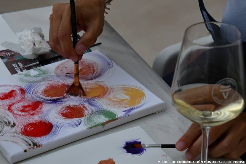 Detalle de una participante pintando una composición abstracta junto a una copa de vino